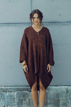 Load image into Gallery viewer, A woman wearing a brown textured knit poncho-style dress stands outdoors against a gray concrete wall, flanked by two tall thin trees.